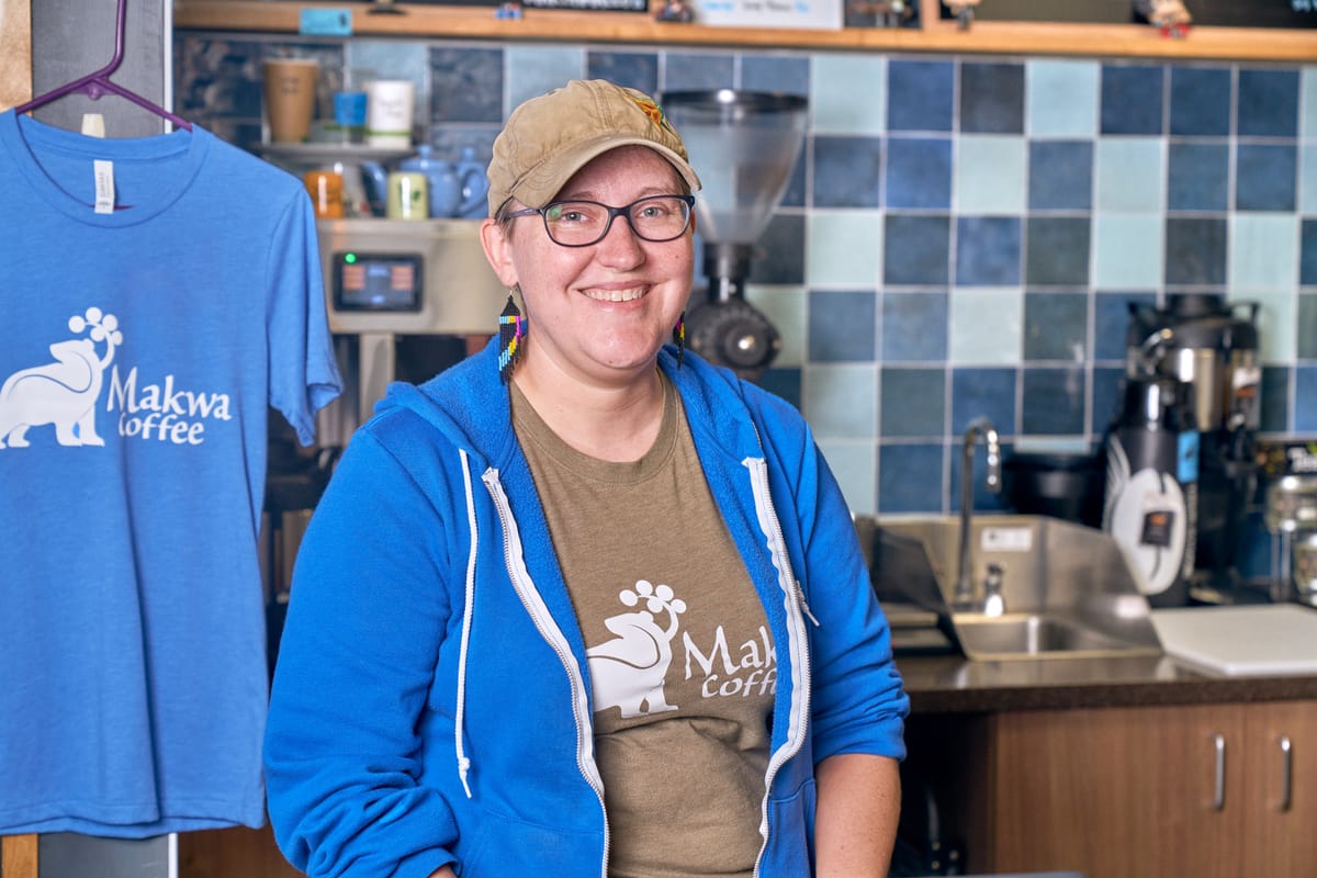 Jamie Becker Finn stands behind the counter at Makwa wearing a bright blue zip up hoodie over a brown Makwa tshirt. The Mawka logo of a bear is on the shirt.