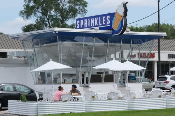 The retro blue and white building has angled glass windows and a blue and white sign on top that says Sprinkles next to an image of an ice cream cone. Two people are sitting at a white table.
