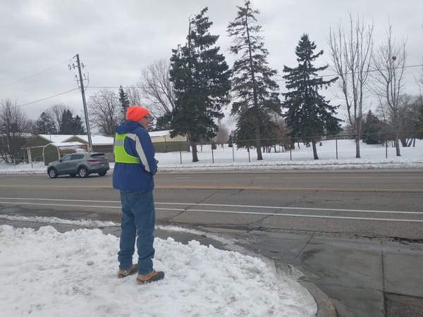 A man wearing a blue winter coat with a bright yellow vest and blaze orange stocking cap stands facing the street on a snowy corner.